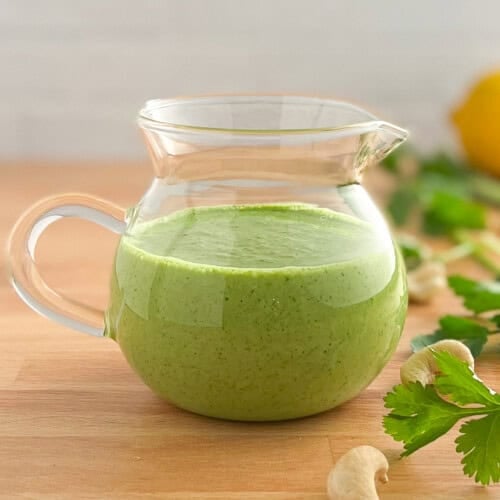 side view close up of green goddess salad dressing in a small glass pitcher with fresh herbs, cashews and lemon blurred in background