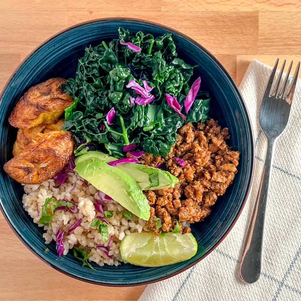 top view close up of sofritas bowl (vegan, oil free) with cilantro brown rice, plantain, steamed kale and sliced avocado in a dark blue bowl sitting on a wood surface