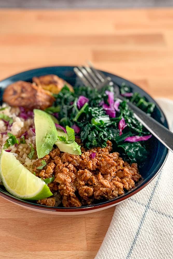 top view close up of sofritas bowl (vegan, oil free) with cilantro brown rice, plantain, steamed kale and sliced avocado in a dark blue bowl sitting on a wood surface