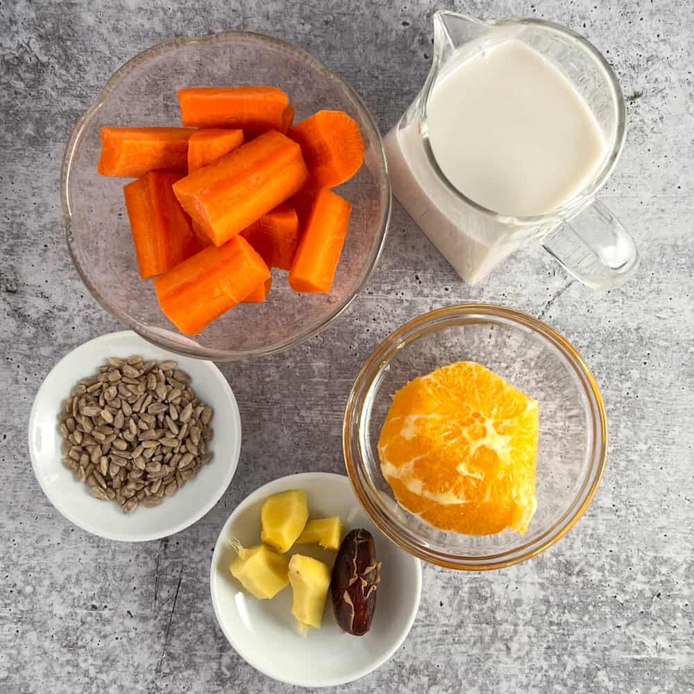 top view of the ingredients for spicy orange creamsicle smoothie on a gray surface. carrot, almond milk, sunflower seeds, ginger root, date, peeled orange
