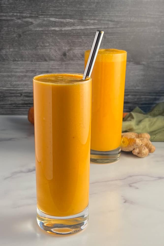 side view of two glasses of the spicy orange creamsicle smoothie with an orange, carrot, ginger and green towel blurred in the background