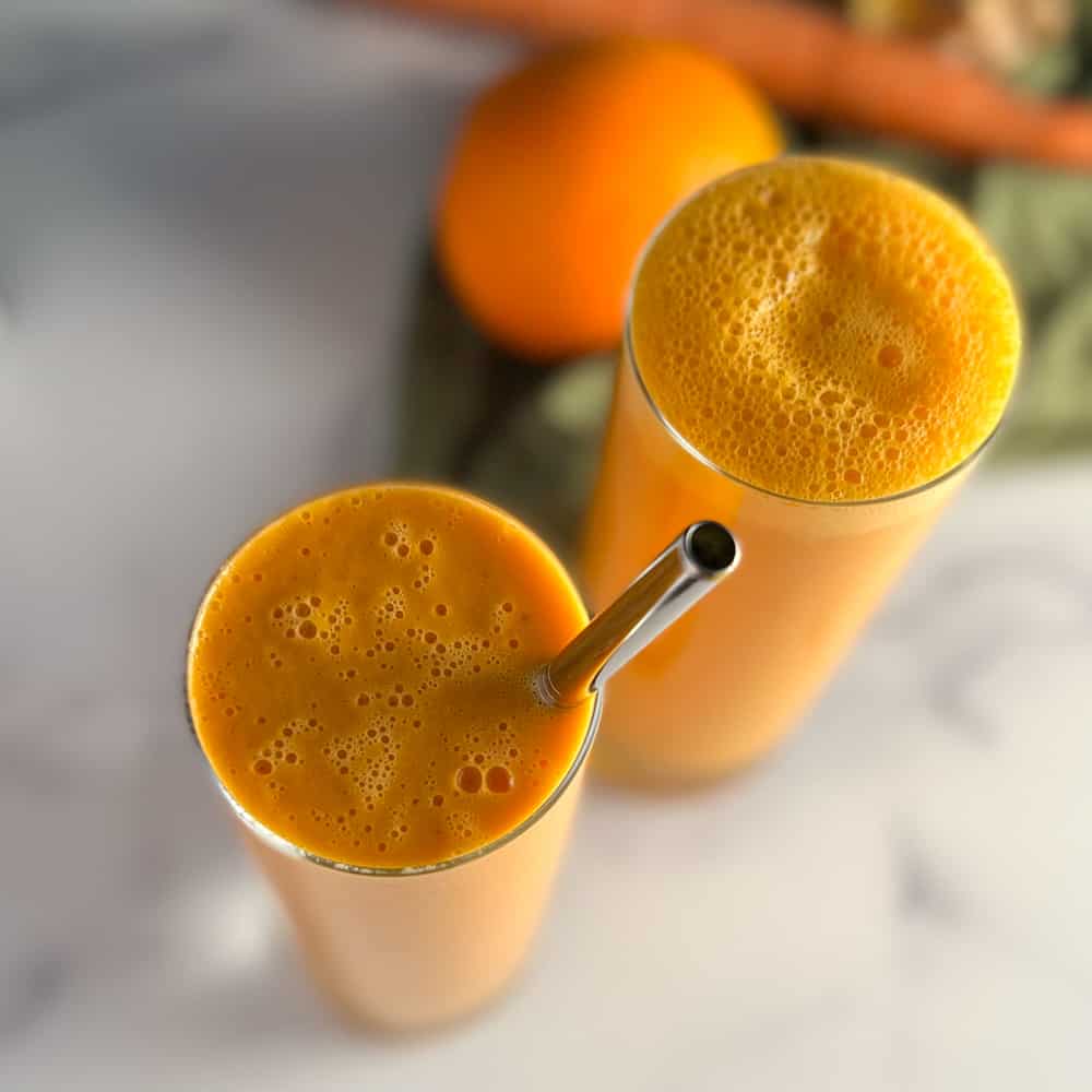 top view of two glasses of the spicy orange creamsicle smoothie with orange, carrot, ginger root and green towel blurred in the background.
