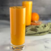 side view of two glasses of spicy orange creamsicle smoothie with orange, carrot, ginger root and green towel blurred in the background