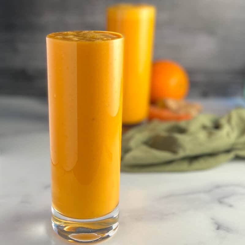 side view of two glasses of spicy orange creamsicle smoothie with orange, carrot, ginger root and green towel blurred in the background