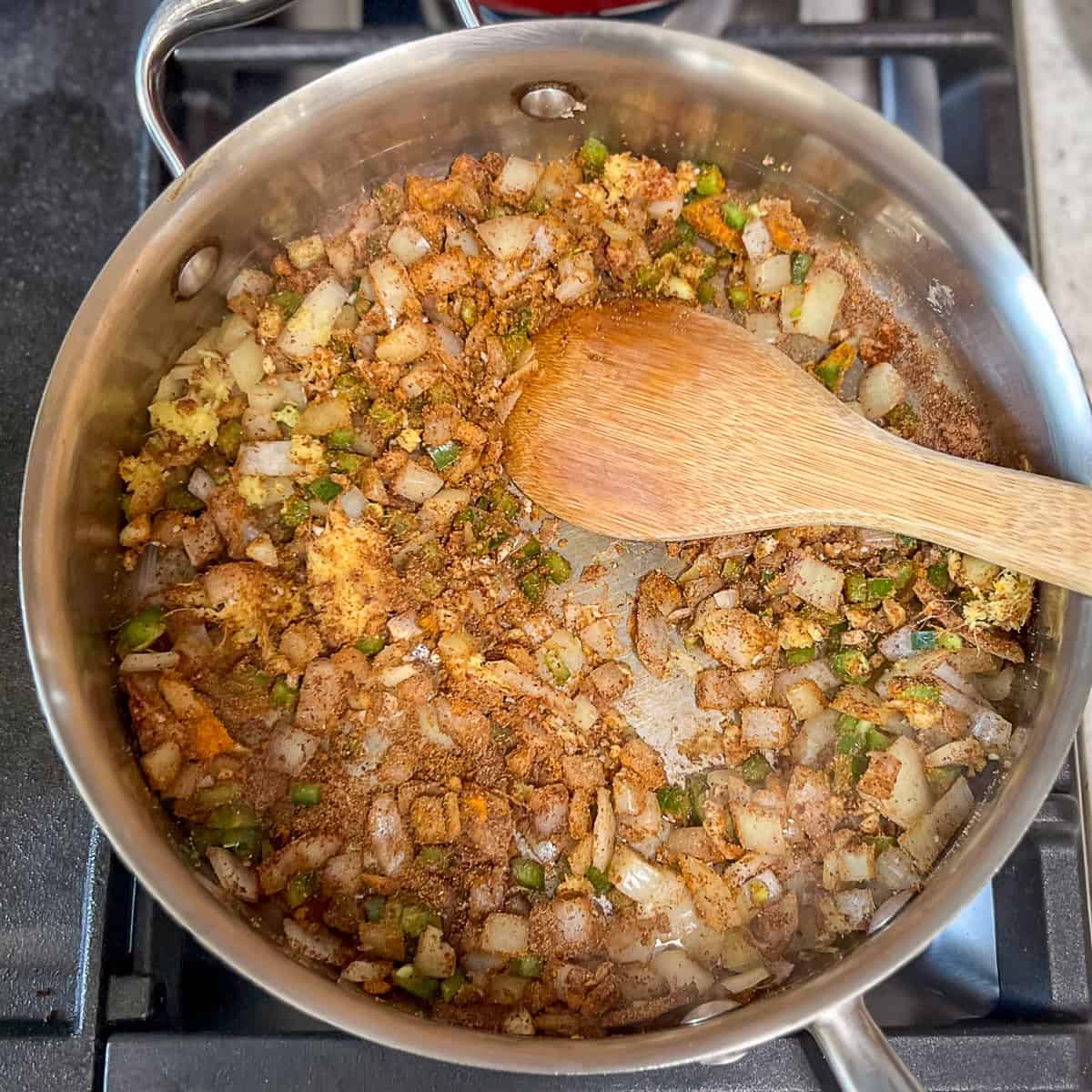 A wooden spoon stirring the aromatics and spices in the skillet.