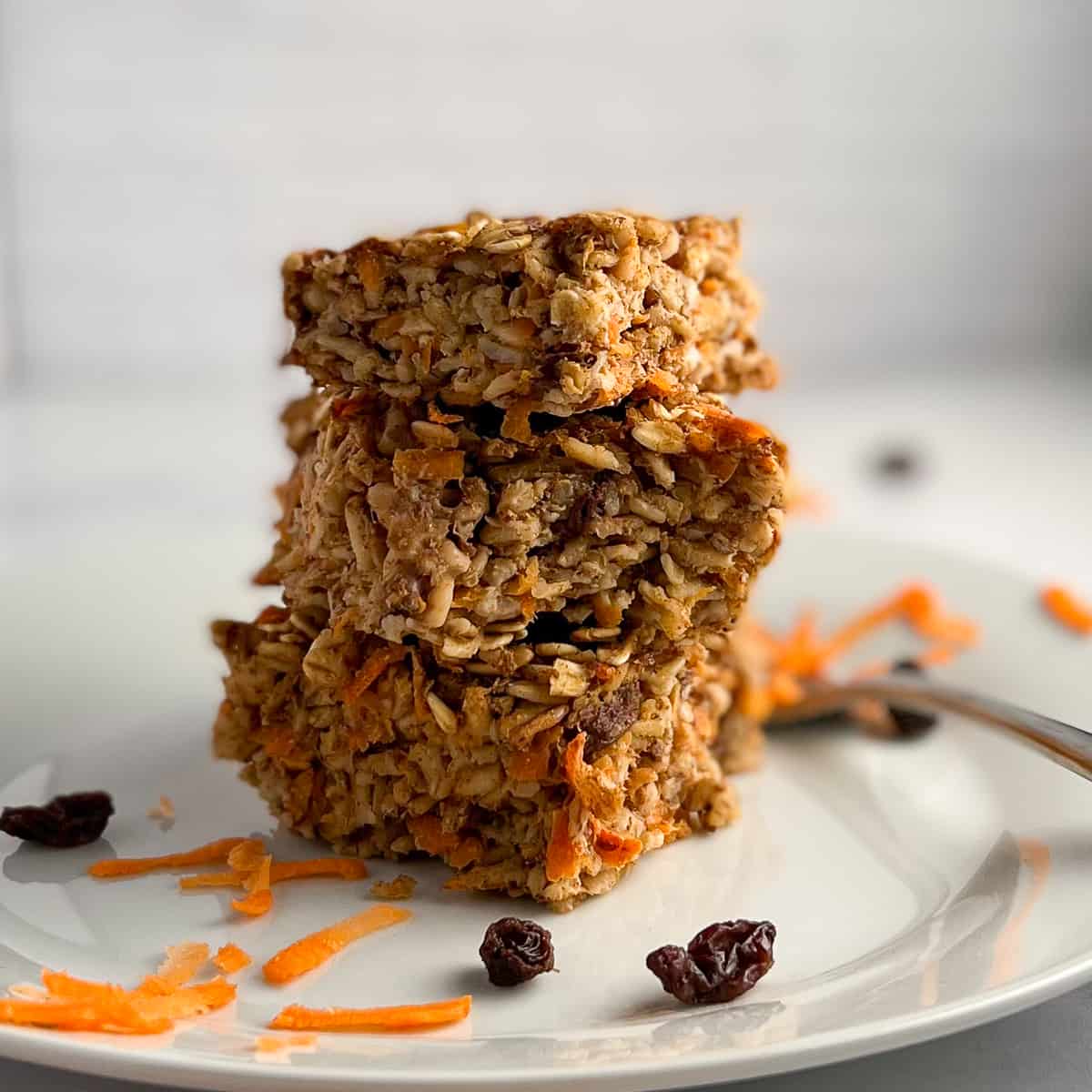 side view close up of carrot cake baked oatmeal on a white plate next to a fork. Shredded carrot and raisins scatter on the plate.