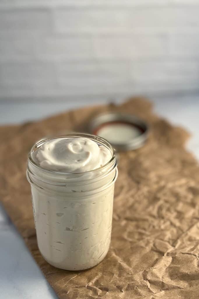 side view of easy vegan mayo in a mason jar with the lid off sitting on crumpled brown paper with the lid blurred in the background