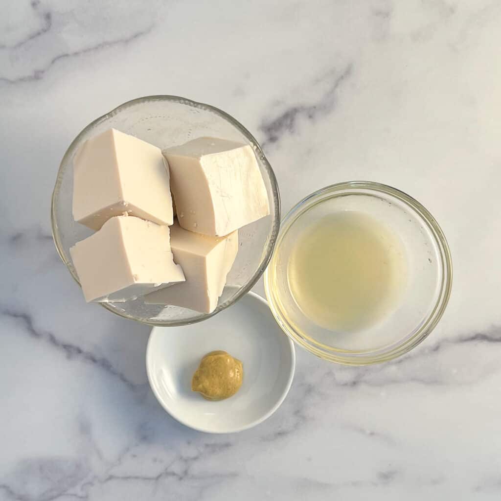 top view of the key ingredients to make easy vegan mayo. silken tofu in large cubes, lemon juice, apple cider vinegar and dijon mustard. the glass and ceramic containers are sitting on a marble surface.