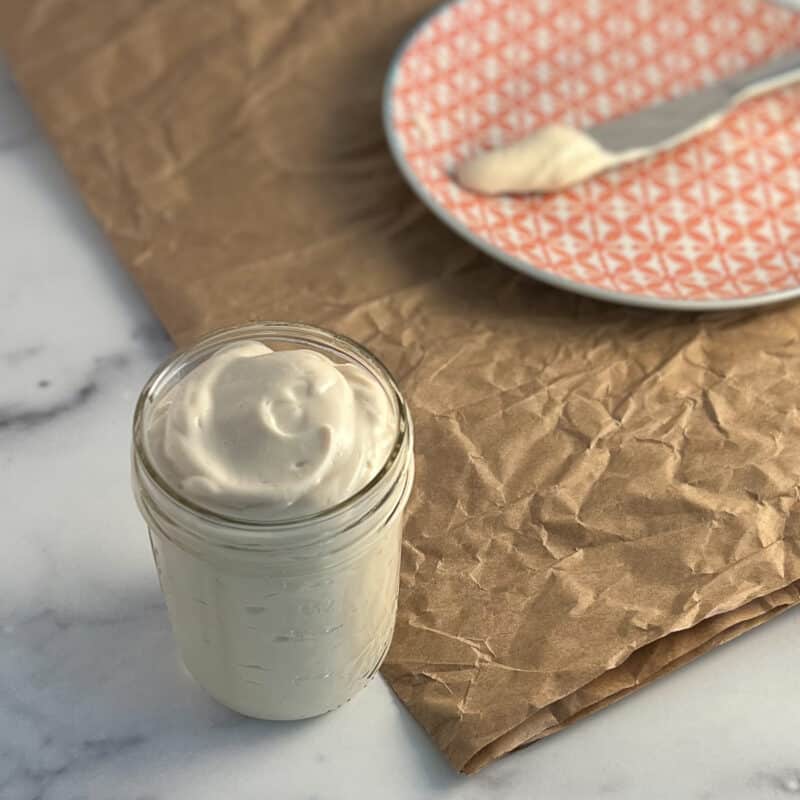 side and top view of easy vegan mayo in a glass mason jar sitting on a marble surface. next to the mayo is a decorative orange and white plate with a knife and some mayo on it sitting on crumbled brown paper