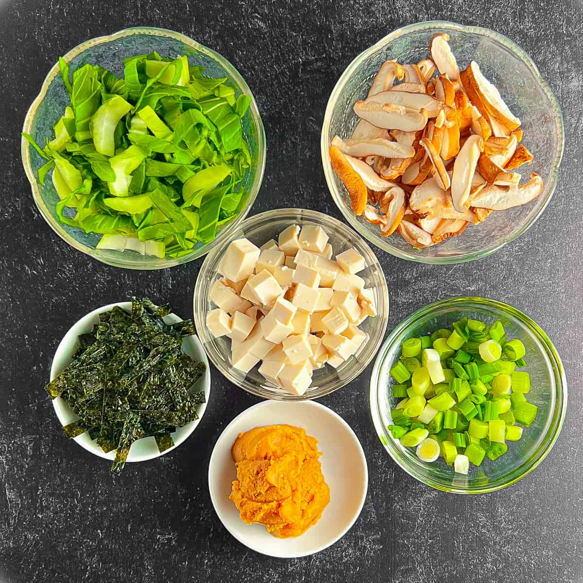 top view of the ingredients for miso soup against a black and gray background: mushrooms, tofu, green onion, miso paste, nori seaweed, baby bok choy