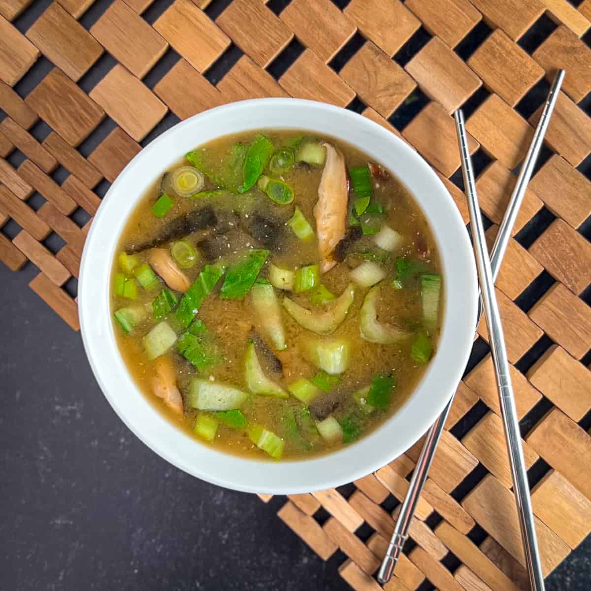 top view close up of miso soup with mushrooms in a white bowl on a wooden placemat next to stainless chopsticks