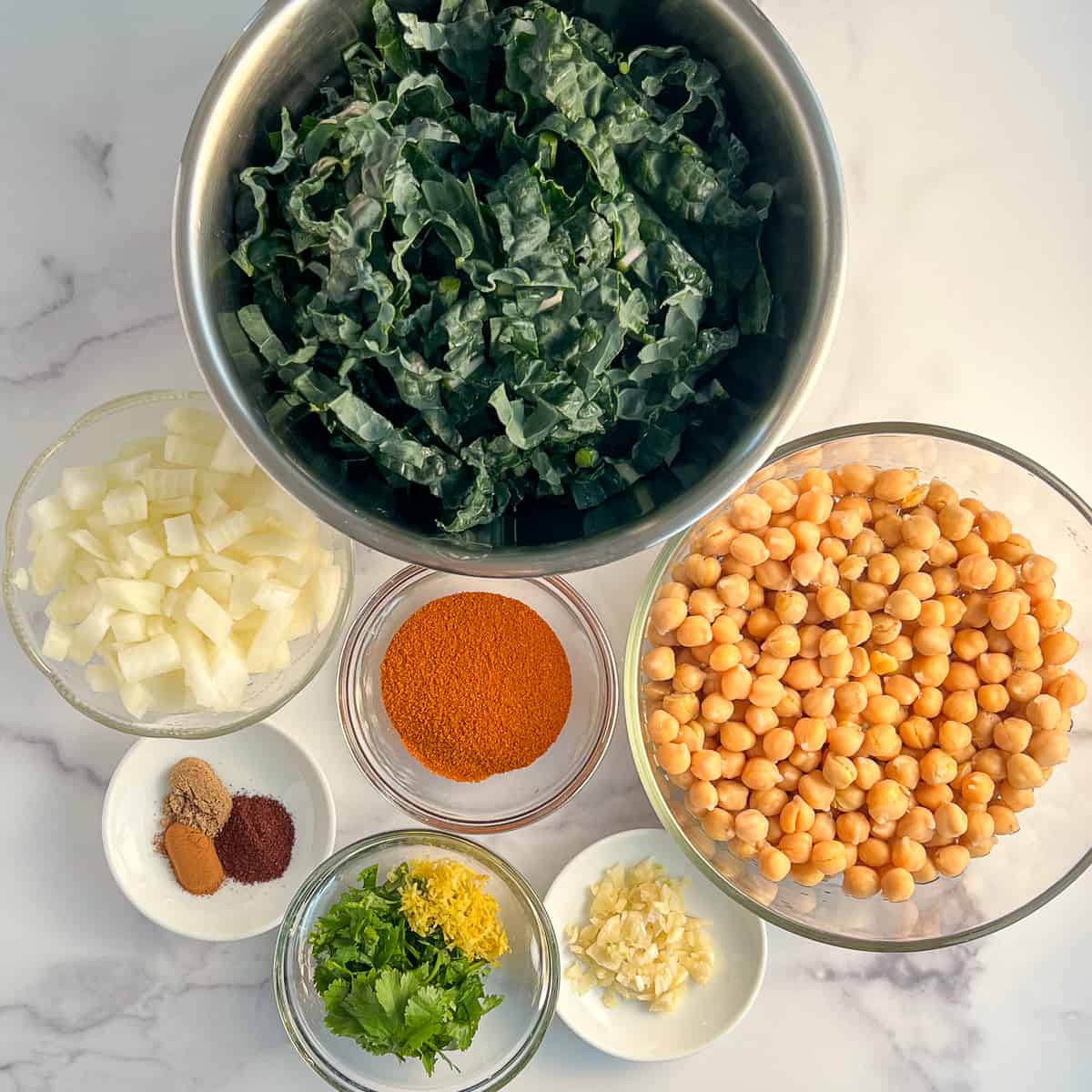 top view of ingredients to make North African Chickpea Soup: kale, onion, chickpeas, harissa, cinnamon, coriander, sumac, garlic, cilantro and lemon zest