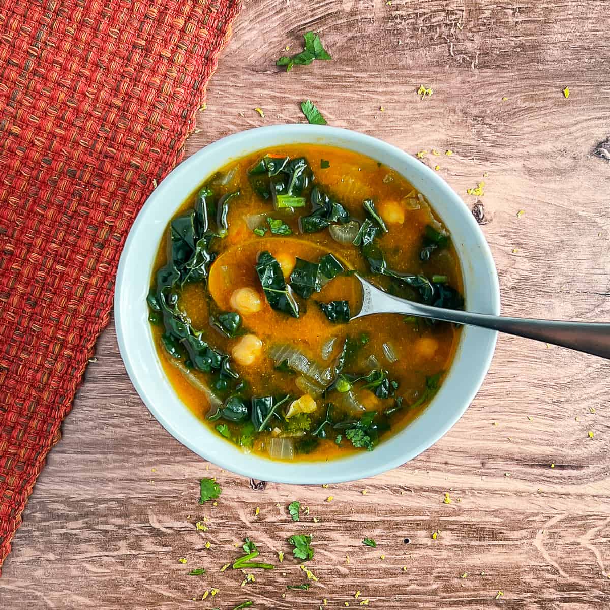 top view close up of North African Chickpea Soup in a white bowl with a spoon on a wooden surface. next to a red placemat