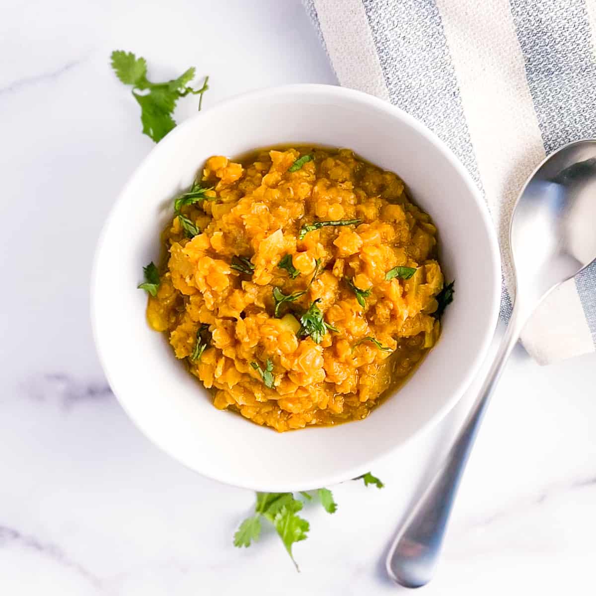 top view close up of red lentil dal in a white bowl on a marble surface with fresh chopped cilantro. spoon and striped blue and white napkin on the side