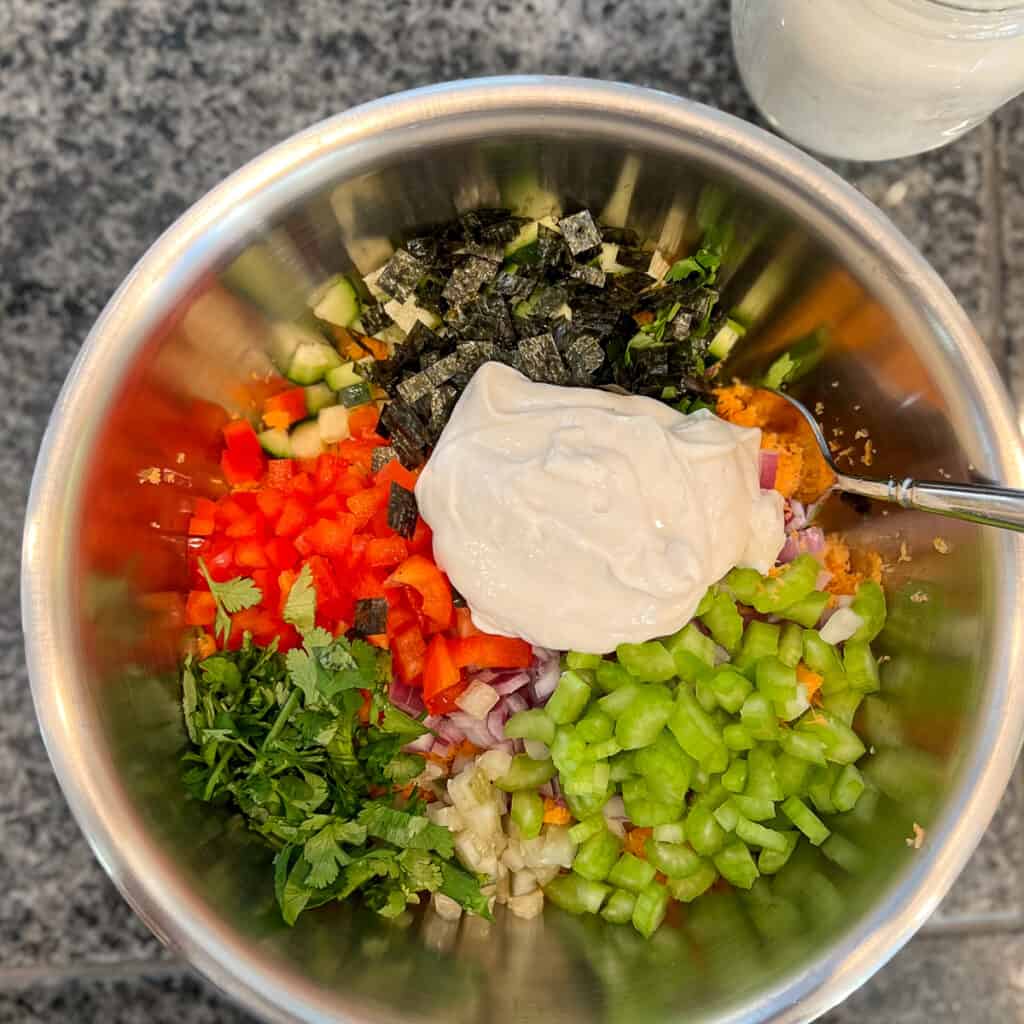 top view of all ingredients-shredded carrots, cilantro, celery, onion, pickle, bell pepper, nori, cucumber and easy vegan may-in a stainless steel mixing bowl with spoon