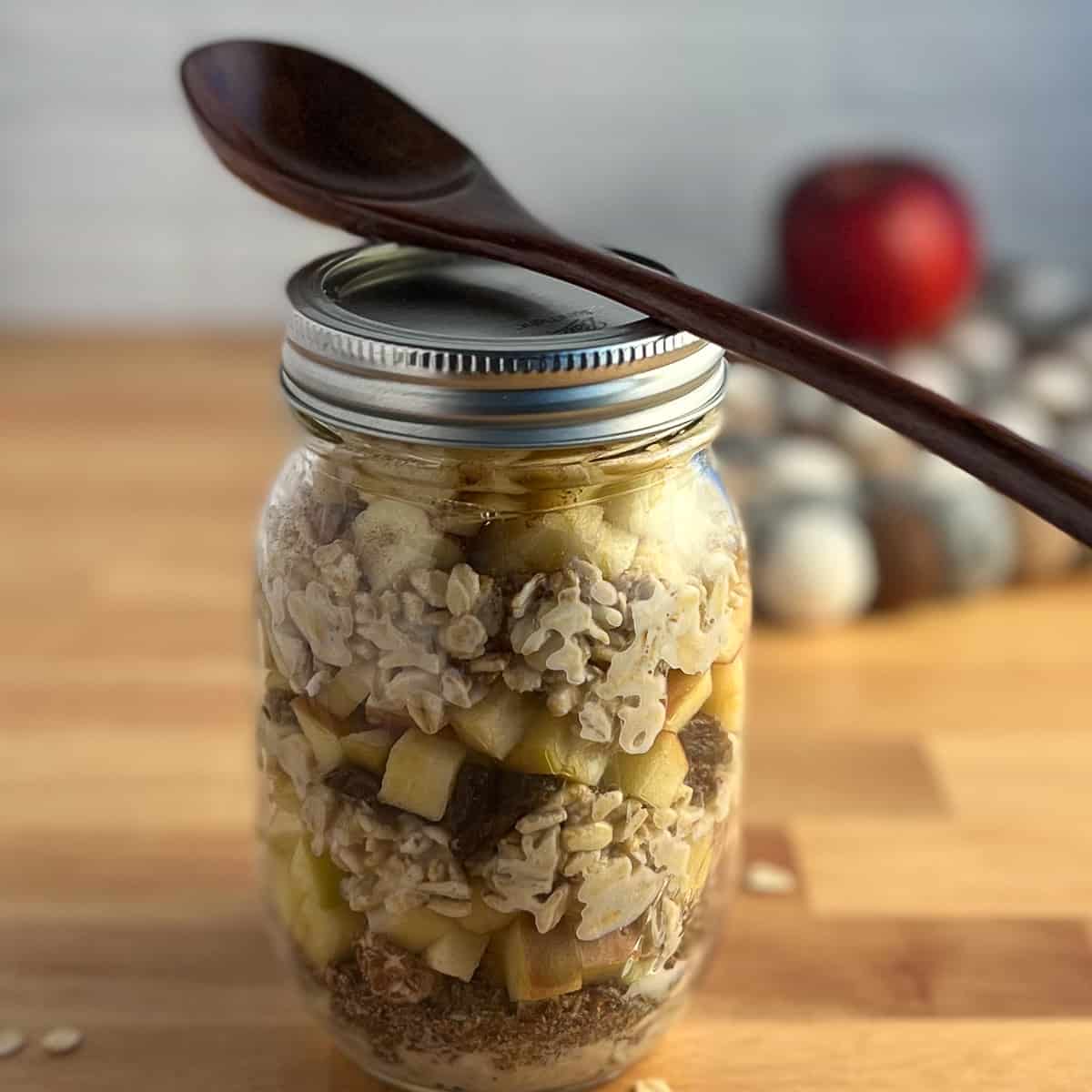 side view of a mason jar with apple cinnamon overnight oats and a wooden spoon balanced on top. red apple blurred in the background
