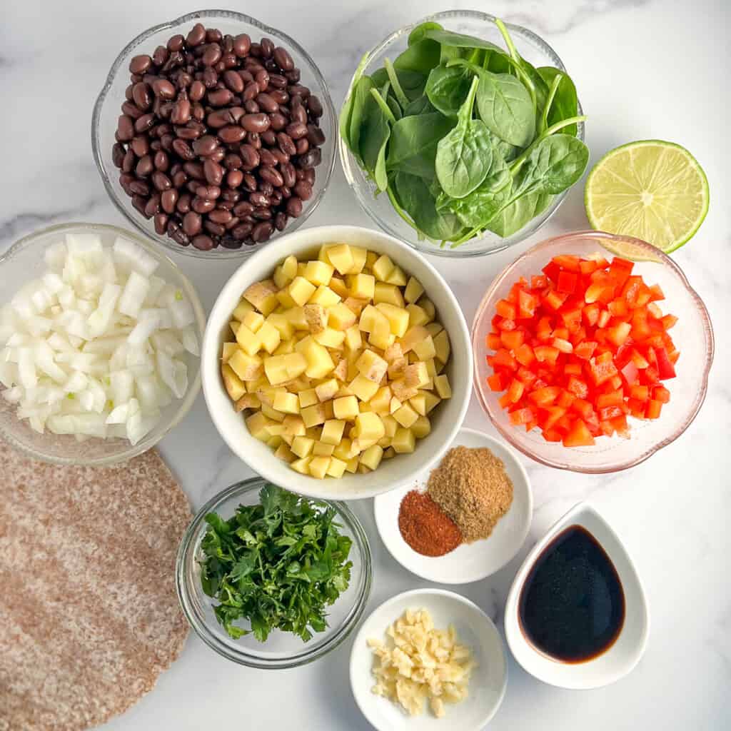 top view of the ingredients used to make hearty vegan breakfast burrito: black beans, spinach, red bell pepper, potatoes, onion, sprouted grain tortillas, cilantro, garlic, coconut aminos, lime and spices