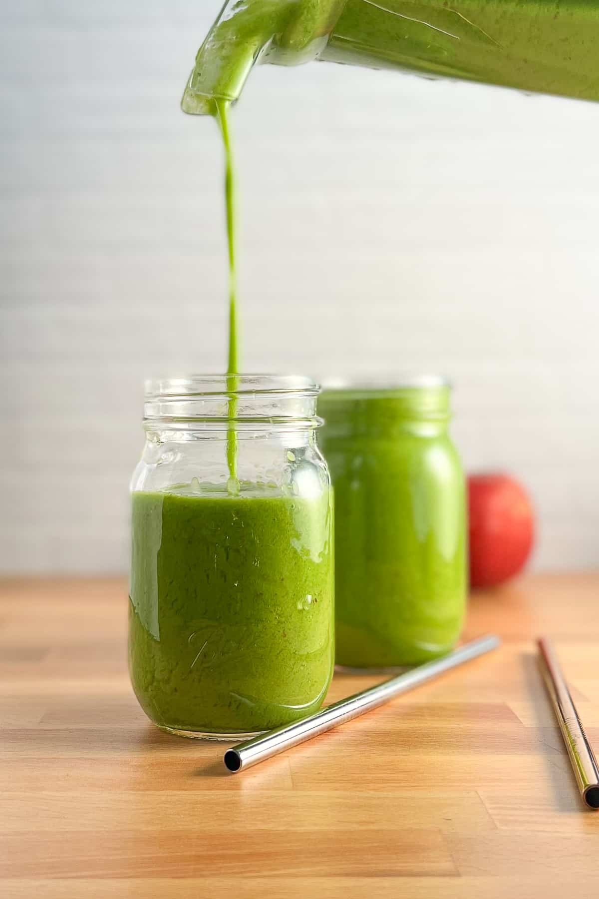 two mason jars with green smoothie being poured into the one of them