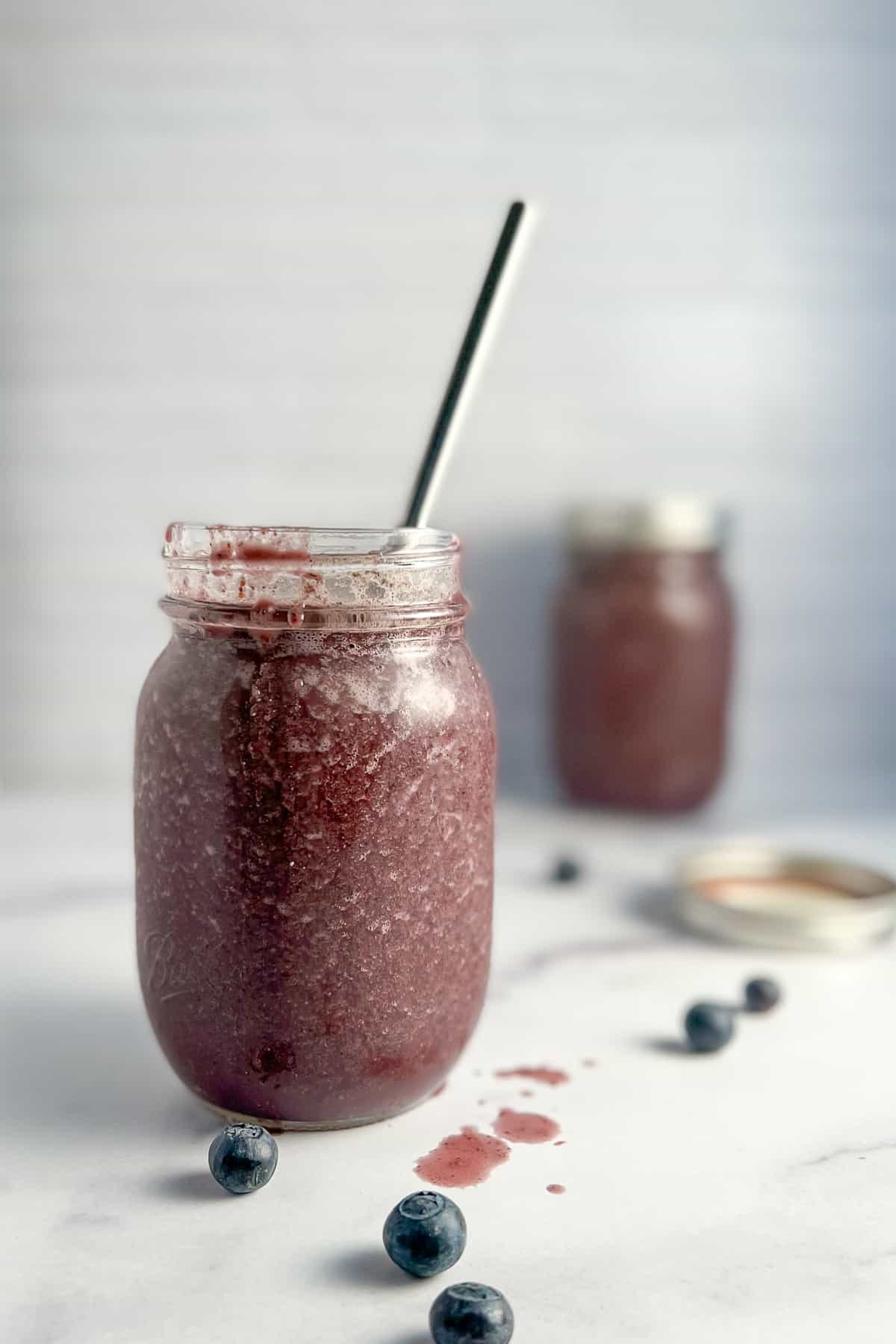 side view of superfood smoothie in a mason jar with a metal straw sticking out of it. a few blueberries are scattered on the surface. a second mason jar with smoothie blurred in the background.