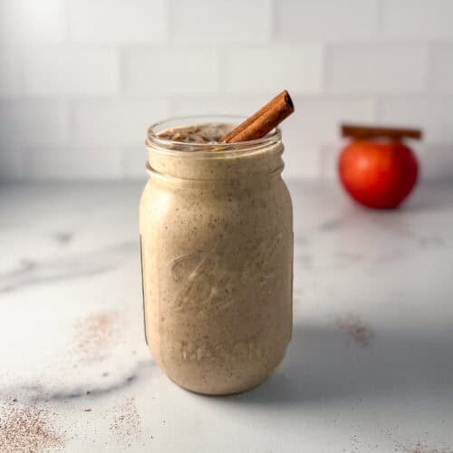 Apple smoothie in a mason jar with a cinnamon stick on the rim; an apple blurred in the background.