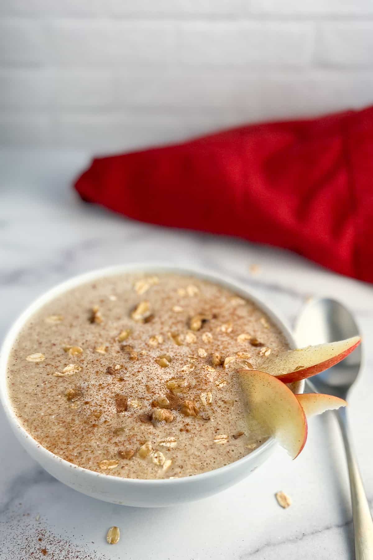 side view of apple smoothie bowl with sliced apples, cinnamon, chopped walnuts and rolled oats on top with spoon and red napkin blurred in the background