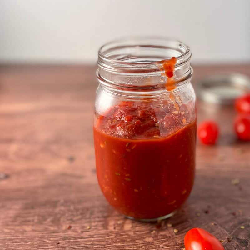 Side view of easy homemade pizza sauce in a glass mason jar on a wood surface with loose grape tomatoes blurred in the background