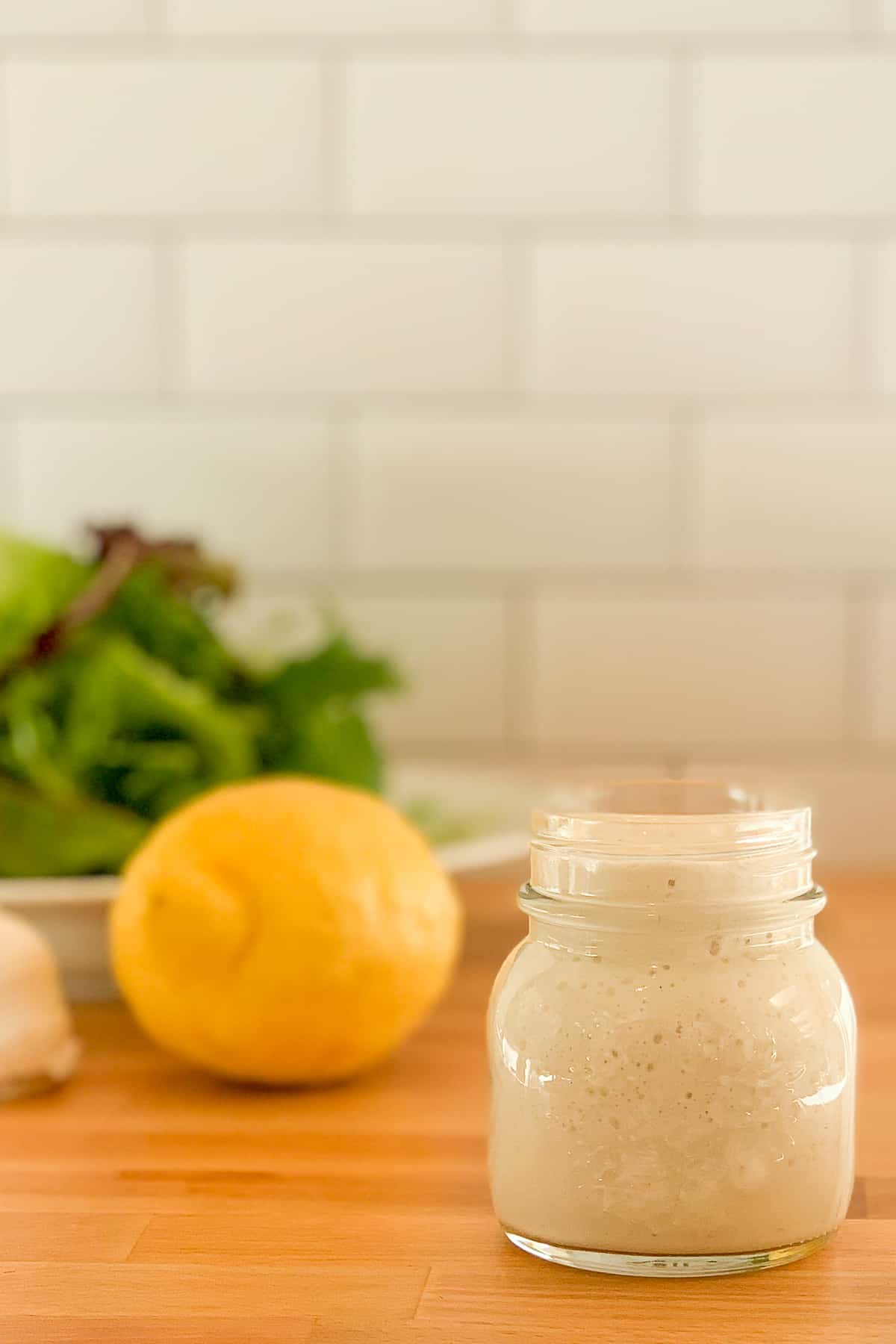 side view close up of tahini lemon garlic sauce in a glass jar with lemon and salad greens blurred in the background