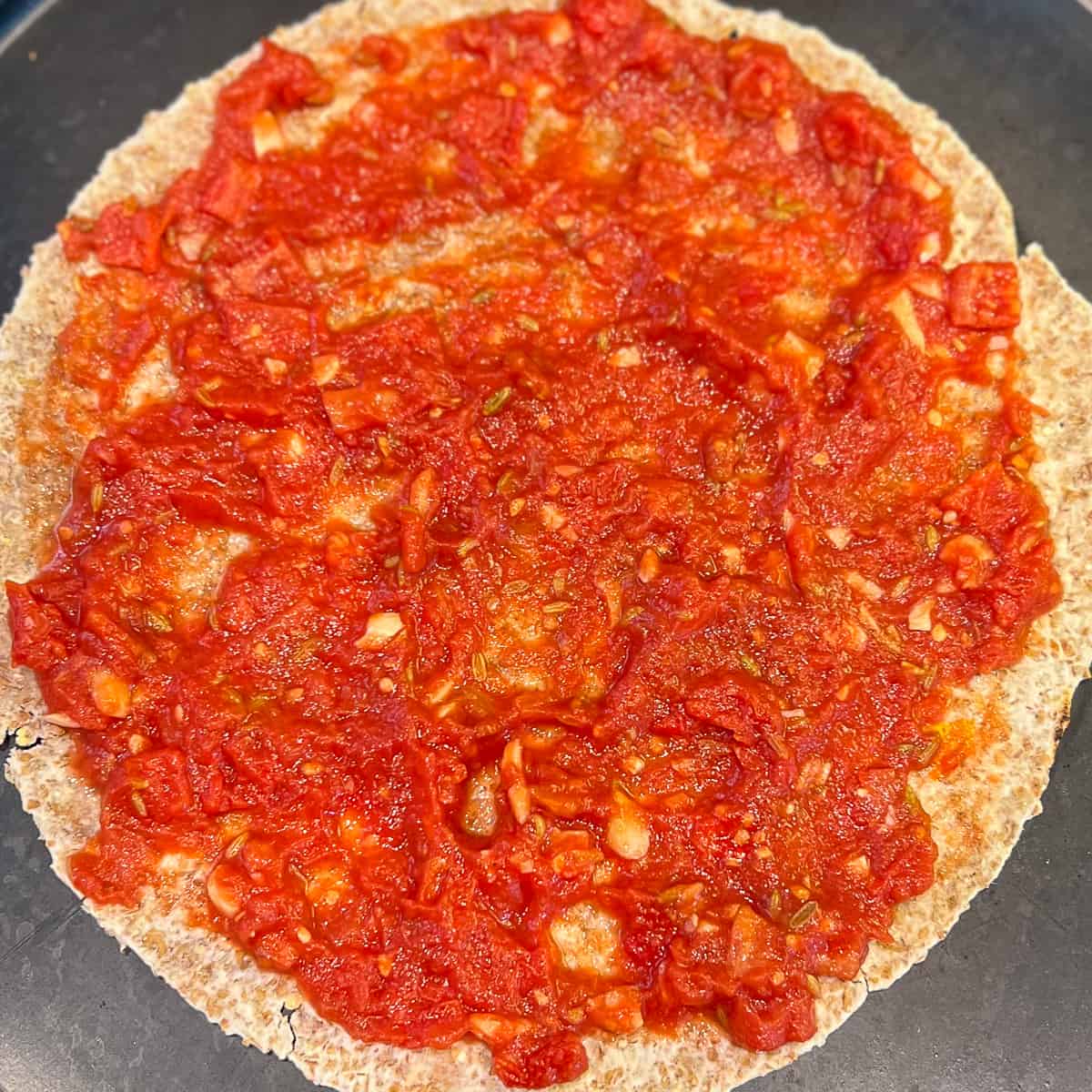 tomato sauce spread on a sprouted grain tortilla being used as the pizza crust