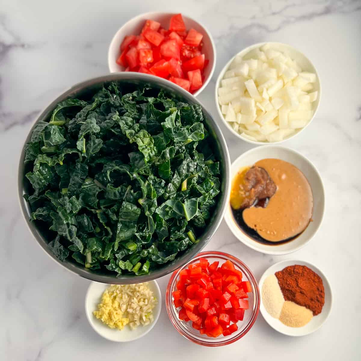 top view of the ingredients for peanut butter greens: kale, tomato, onion, garlic, ginger, bell pepper, peanut butter, Simple Homemade Date Syrup, Scotch Bonnet pepper sauce, coconut aminos, smoked paprika, garlic powder, onion powder