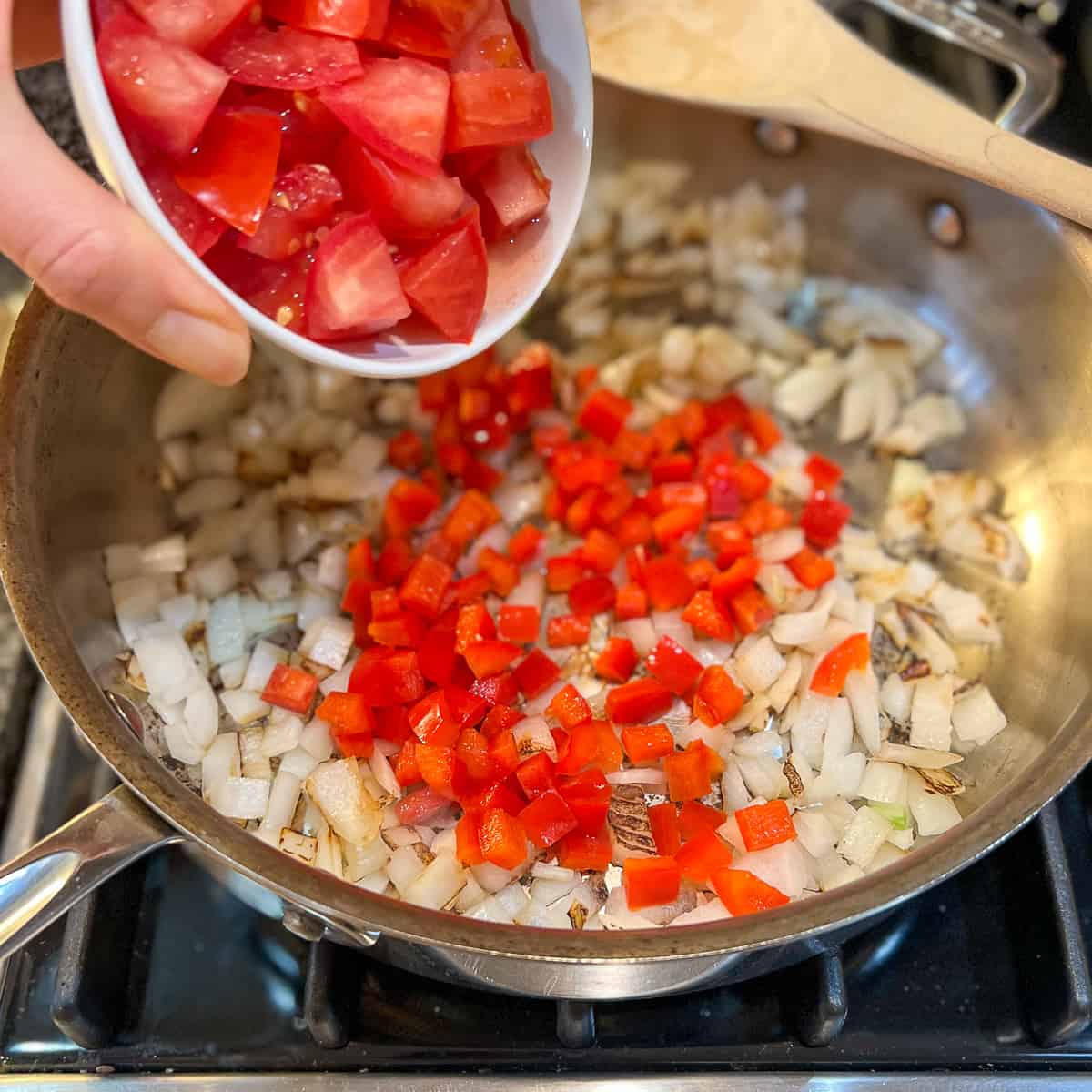 Chopped tomatoes being added to a skillet with bell pepper and onions.