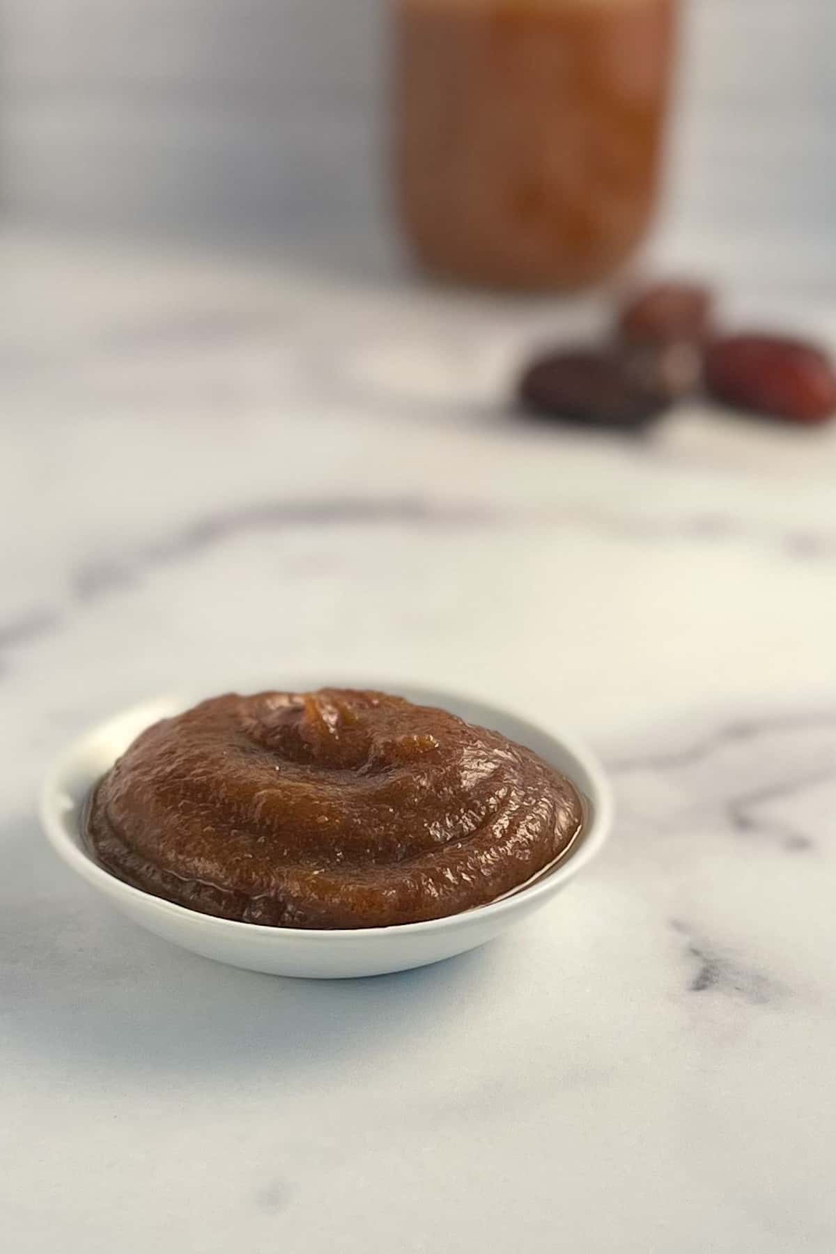 side view close up of simple homemade date syrup in a small white bowl sitting on a marble surface, with dates and a container of syrup blurred in the background