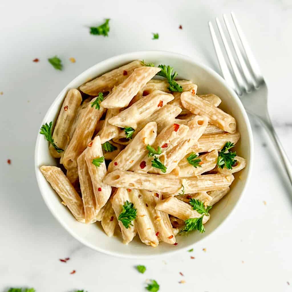 Top view close up of simple tahini pasta in a white bowl with fresh parsley and crushed red pepper next to a fork 