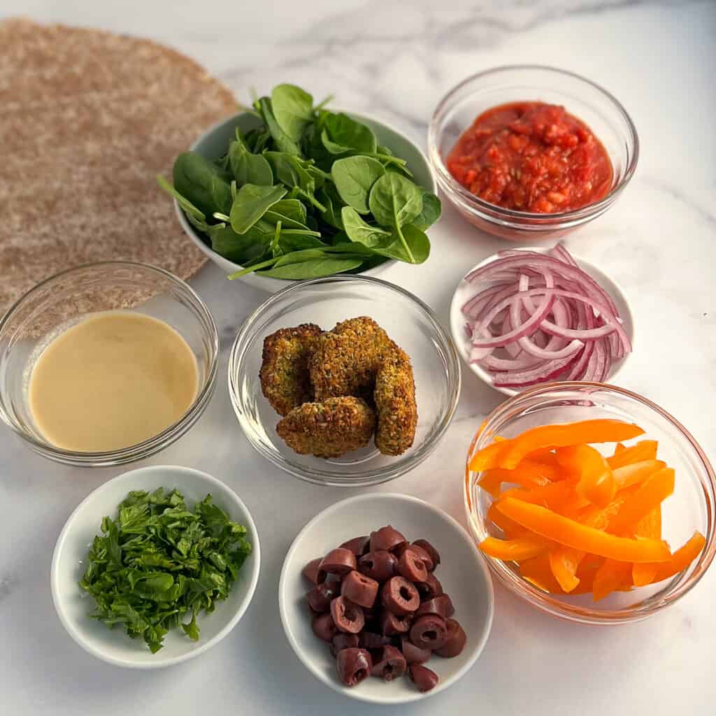 top view of key ingredients: Ezekiel sprouted grain tortilla, spinach, homemade pizza sauce, falafel, onion, bell pepper, olives, parsley and epic tahini lemon garlic sauce