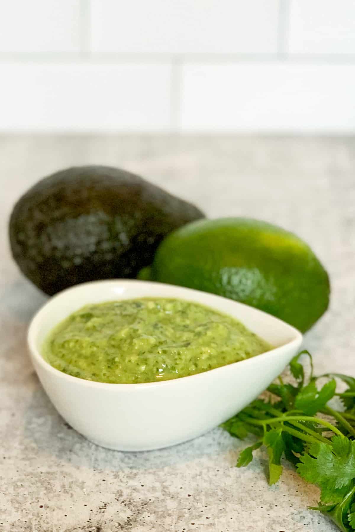 a small white ceramic bowl of avocado cilantro lime dressing next to an avocado, lime and springs of fresh cilantro