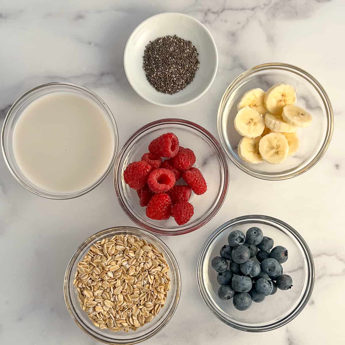 Top view of the key ingredients for overnight oats: rolled oats, almond milk, raspberries, blueberries, banana, chia seeds