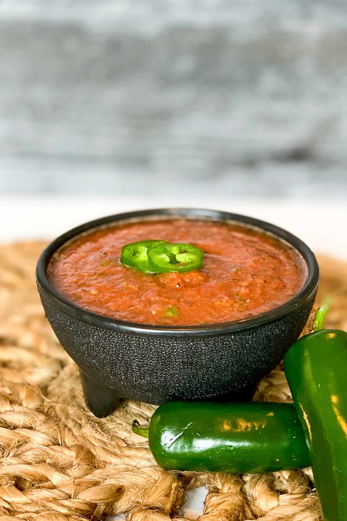 side top view of salsa in a black bowl with sliced jalapeno on top and whole jalapenos on the side