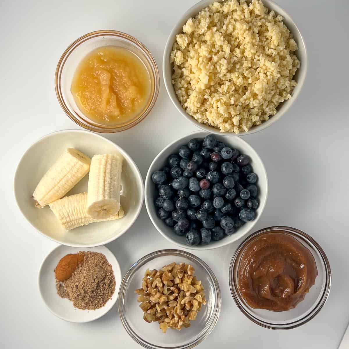 top view of the ingredients for flourless millet muffins: unsweetened apple sauce, millet (cook and cooled), banana, fresh blueberries, ground flaxseeds, cinnamon, cardamom, simple homemade date syrup, chopped walnuts