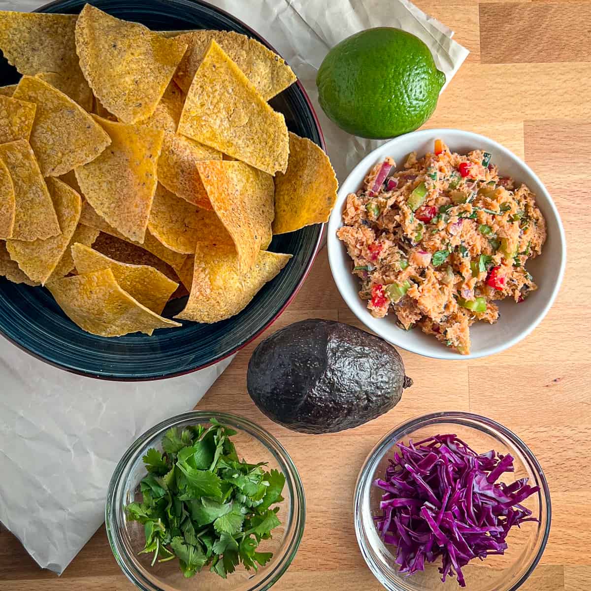 top view of the ingredients for deconstructed vegan fish tacos: oven baked tortilla chips, lime, vegan tuna salad, avocado, fresh cilantro and purple cabbage