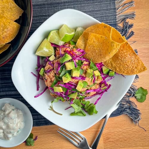 top view close of a bowl of deconstructed vegan fish tacos with vegan tuna salad topped with cabbage, avocado and cilantro. fresh lime, corn tortillas and vegan fish taco sauce on the side.