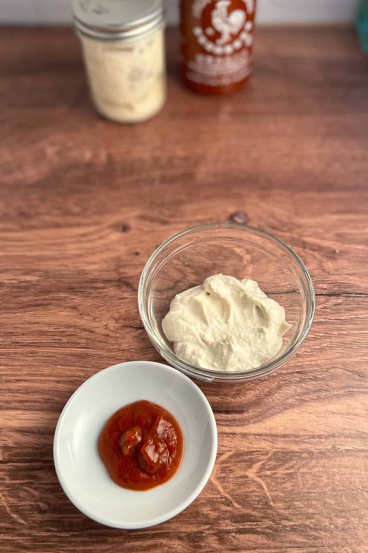 top side view of vegan mayo and sriracha against a wooden surface. mason jar with mayo and bottle of sriracha chili sauce blurred in the background.