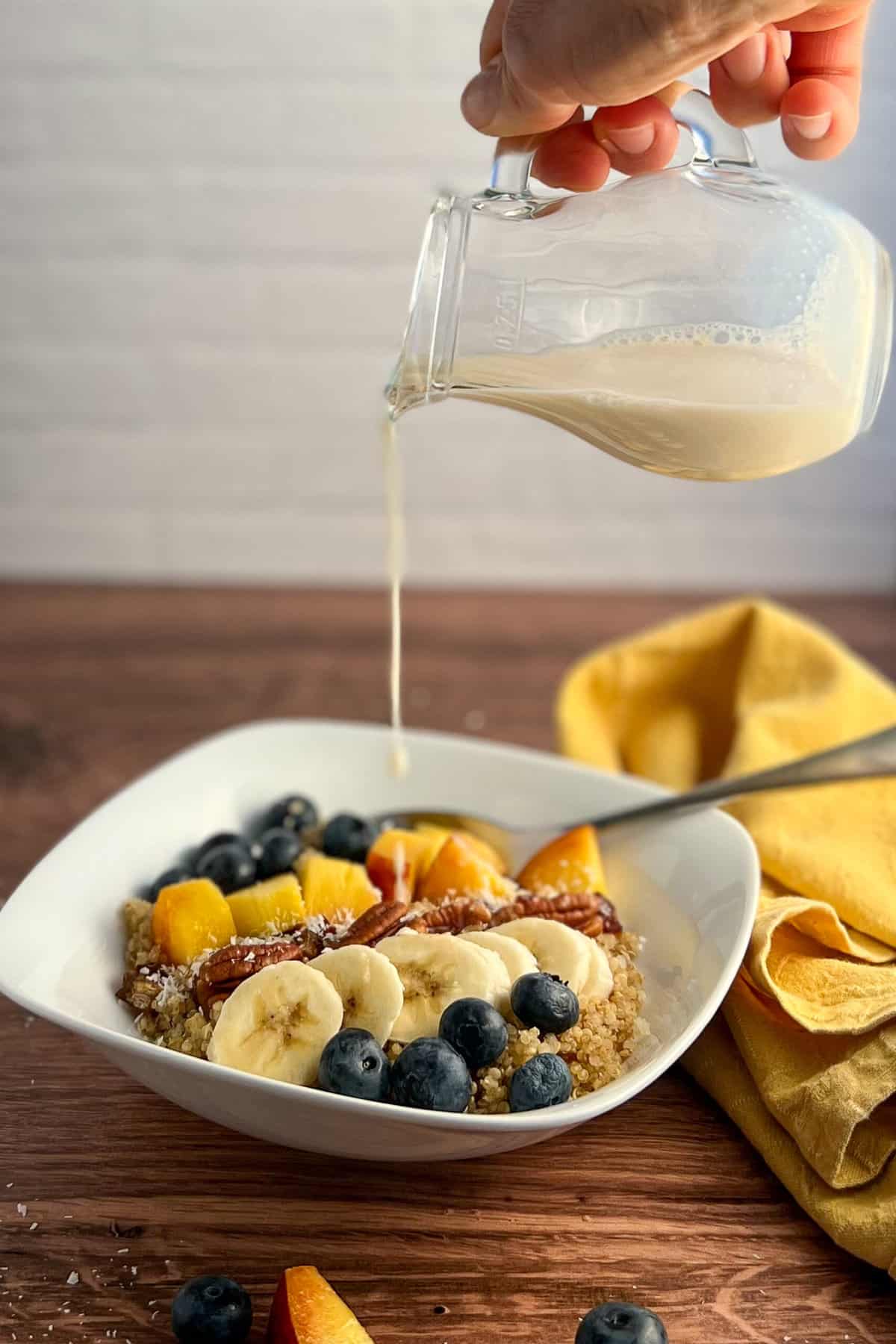 side view of vegan quinoa breakfast bowl in a square white ceramic bowl with a spoon and napkin blurred on the side. a woman's hand is holding a small glass pitcher of plant based milk being poured into the bowl.