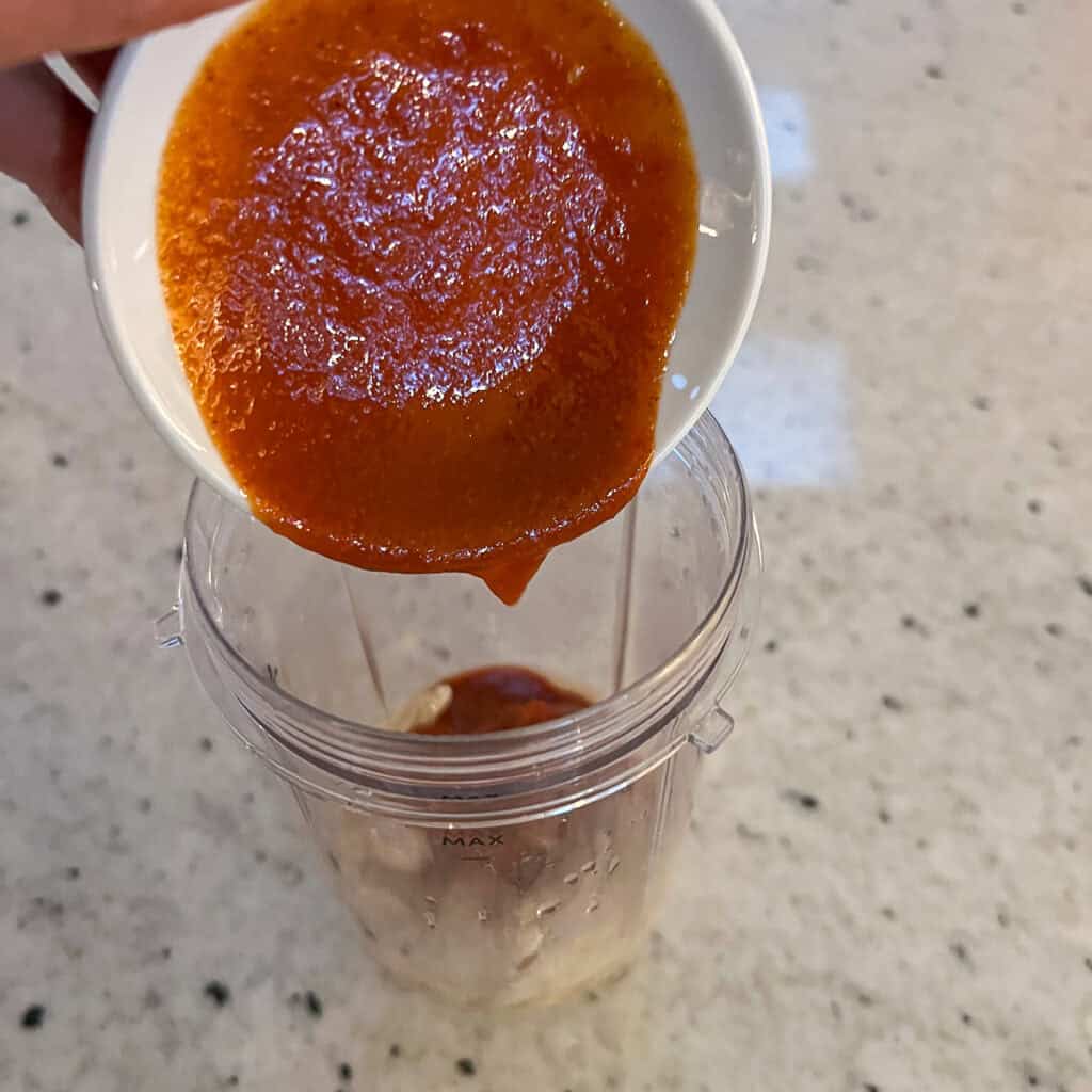Sriracha being added to a blender cup with the other ingredients.