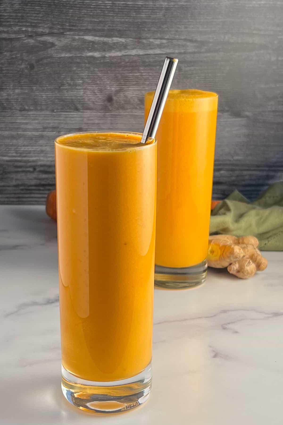 side view of two glasses of the spicy orange creamsicle smoothie with an orange, carrot, ginger and green towel blurred in the background