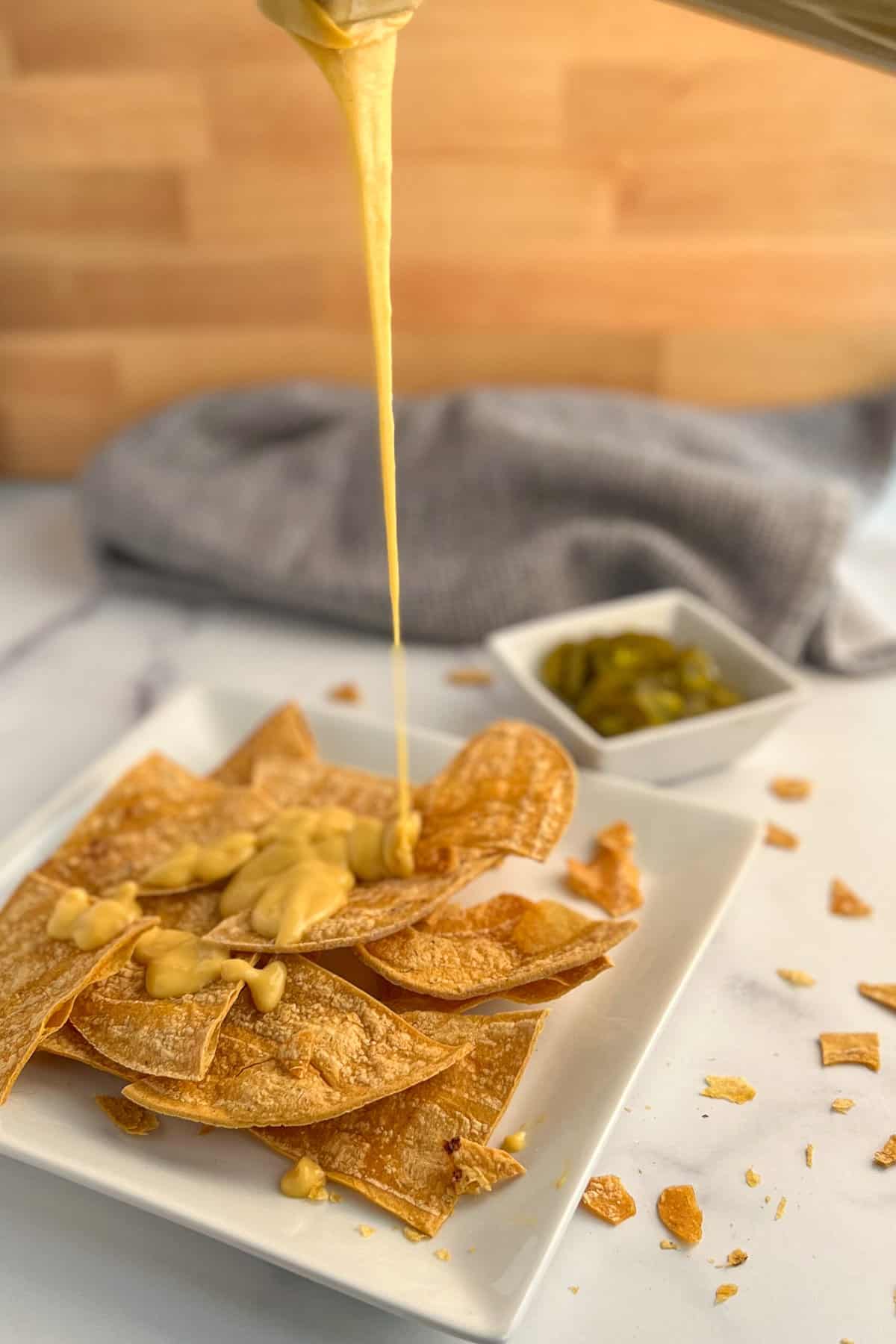 side view of vegan cheese sauce being poured onto oven baked tortilla chips on a square white plate. pickled jalapenos and gray dish towel blurred in the background