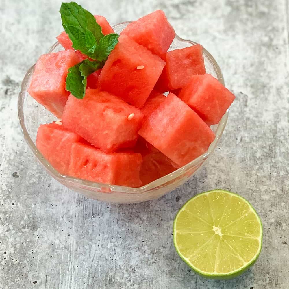 top side view of watermelon cubes in a glass bowl with fresh mint sprig on top and ½ lime on the side