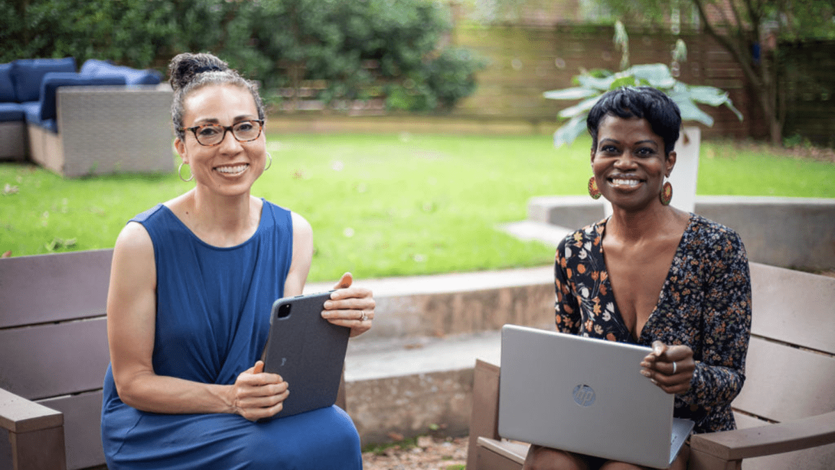two women sitting outside smiling at the camera holding a laptop and tablet.