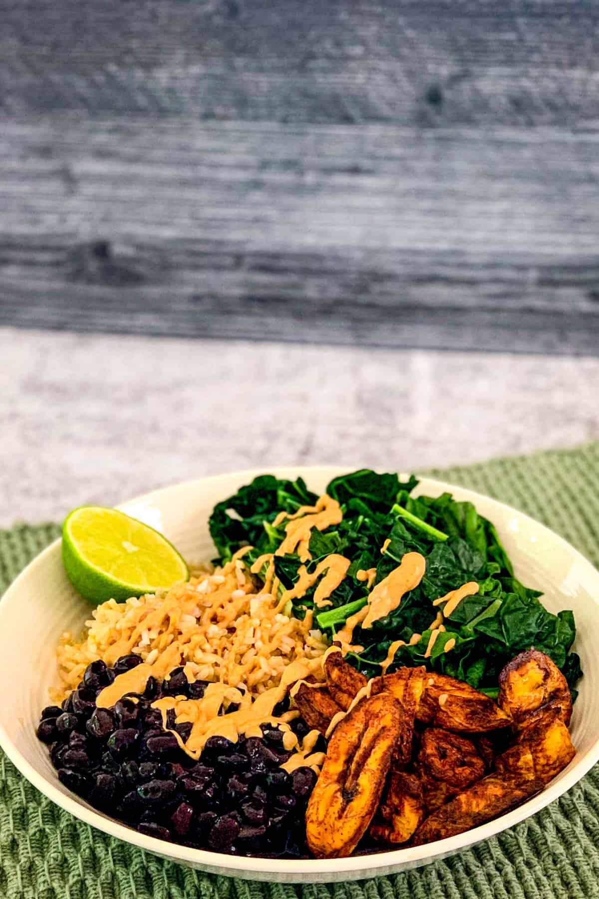 top side view of black bean buddha bowl with black beans, plantains, kale, brown rice and lime wedge in a white bowl on a green placemat with gray background.