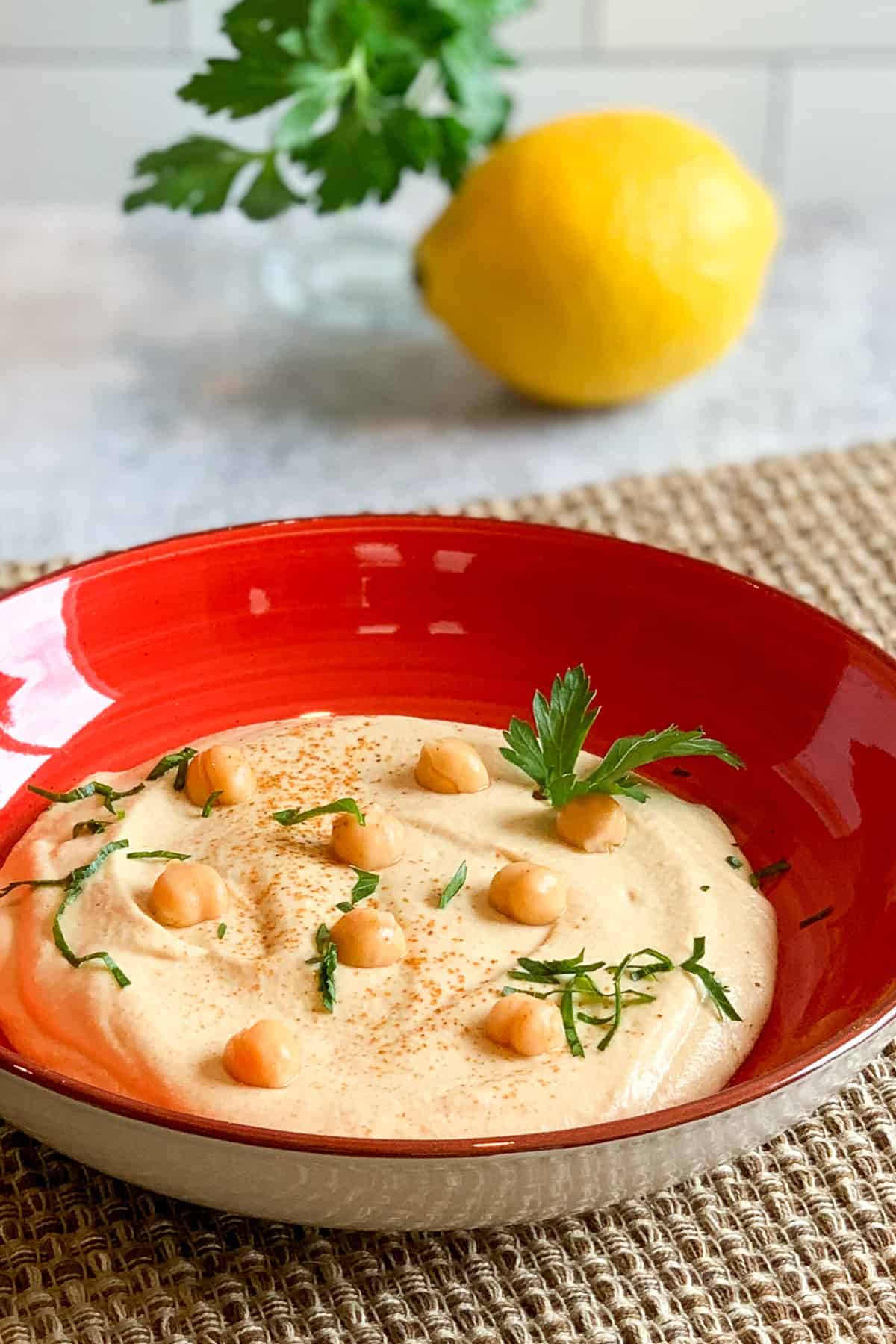 Best Garlic Hummus in a red bowl on a woven mat with fresh parsley on top and blurred in the background next to a lemon