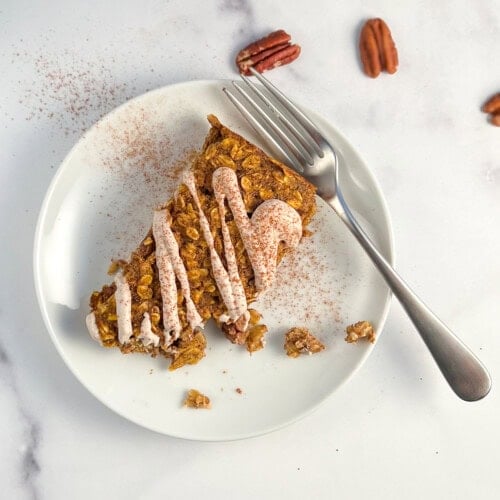 top view close up of a pumpkin oatmeal bar with sweet cashew cream drizzled on top on a round white plate, and fork on the side. pumpkin pie spice dusted on top with loose pecans on the side