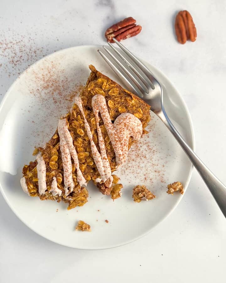 top view close up of a pumpkin oatmeal bar with sweet cashew cream drizzled on top on a round white plate, and fork on the side. pumpkin pie spice dusted on top with loose pecans on the side
